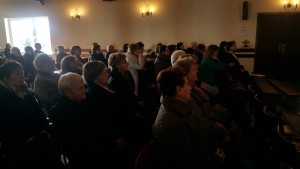 Grupa osób uczestniczy w Dniu Skupienia Wolontariatu Hospicyjnego w sali z oknami i lampami ściennymi.
