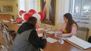 Dwie osoby siedzą przy stole podczas spotkania, w tle widać obraz religijny i balony.