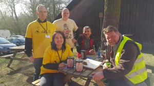 Wolontariusze w żółtych koszulkach i kamizelce, siedzą przy piknikowym stole z jedzeniem podczas zbiórki.