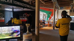 Nagrody sportowe stoją na stole, a osoba w żółtej koszulce filmuje wydarzenie w hali.