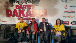 Czterech mężczyzn i kobieta siedzą na krzesłach przed banerem z logotypami sponsorów rajdu Dakar.