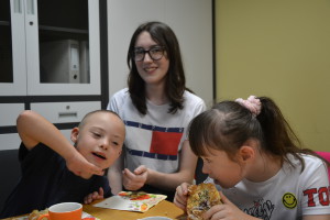 Uczestnicy warsztatów spożywają posiłek przy wsparciu osoby dorosłej w jasnym pomieszczeniu.