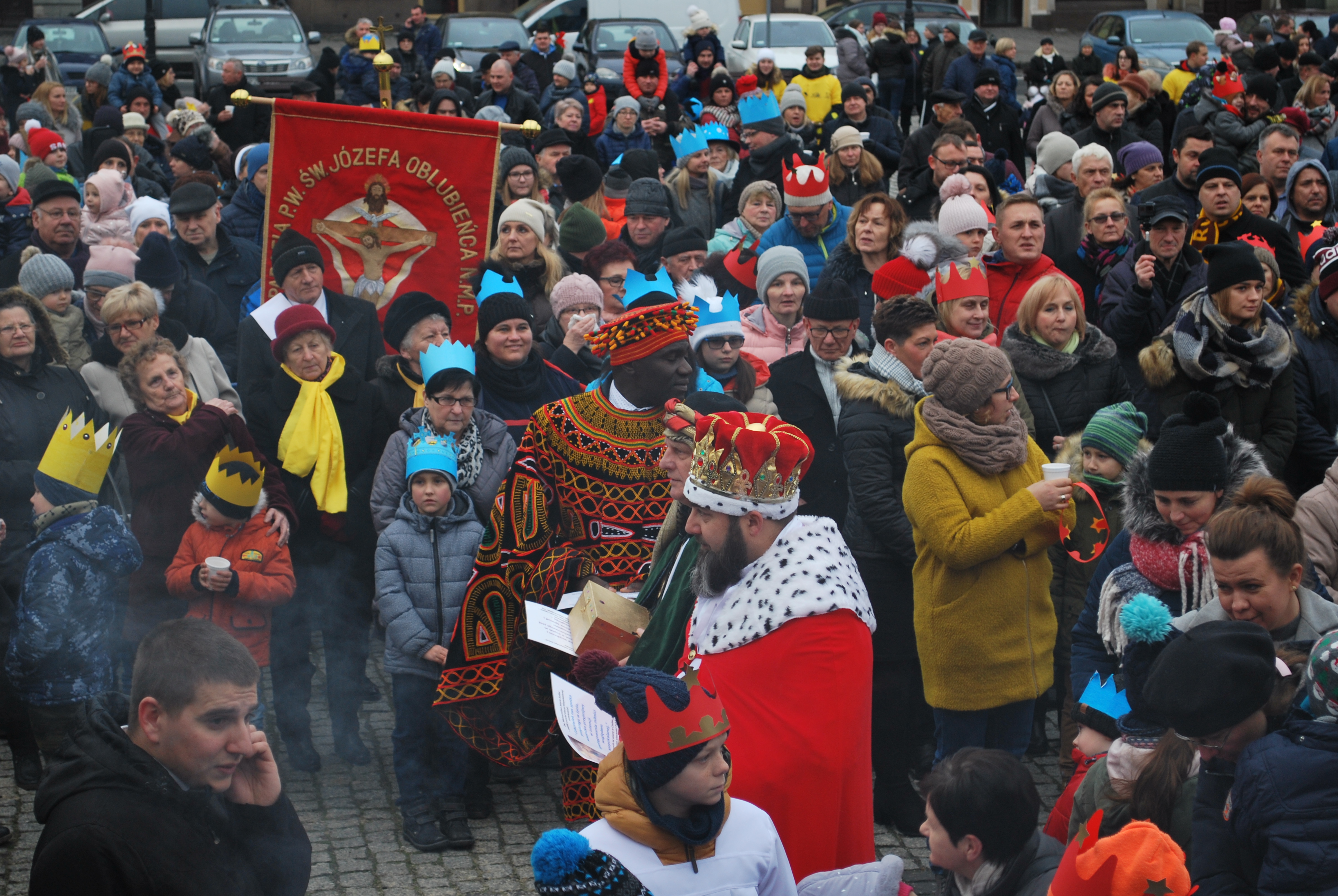 Korowód Trzech Króli w tłumie ludzi, z flagą i uczestnikami w barwnych strojach.