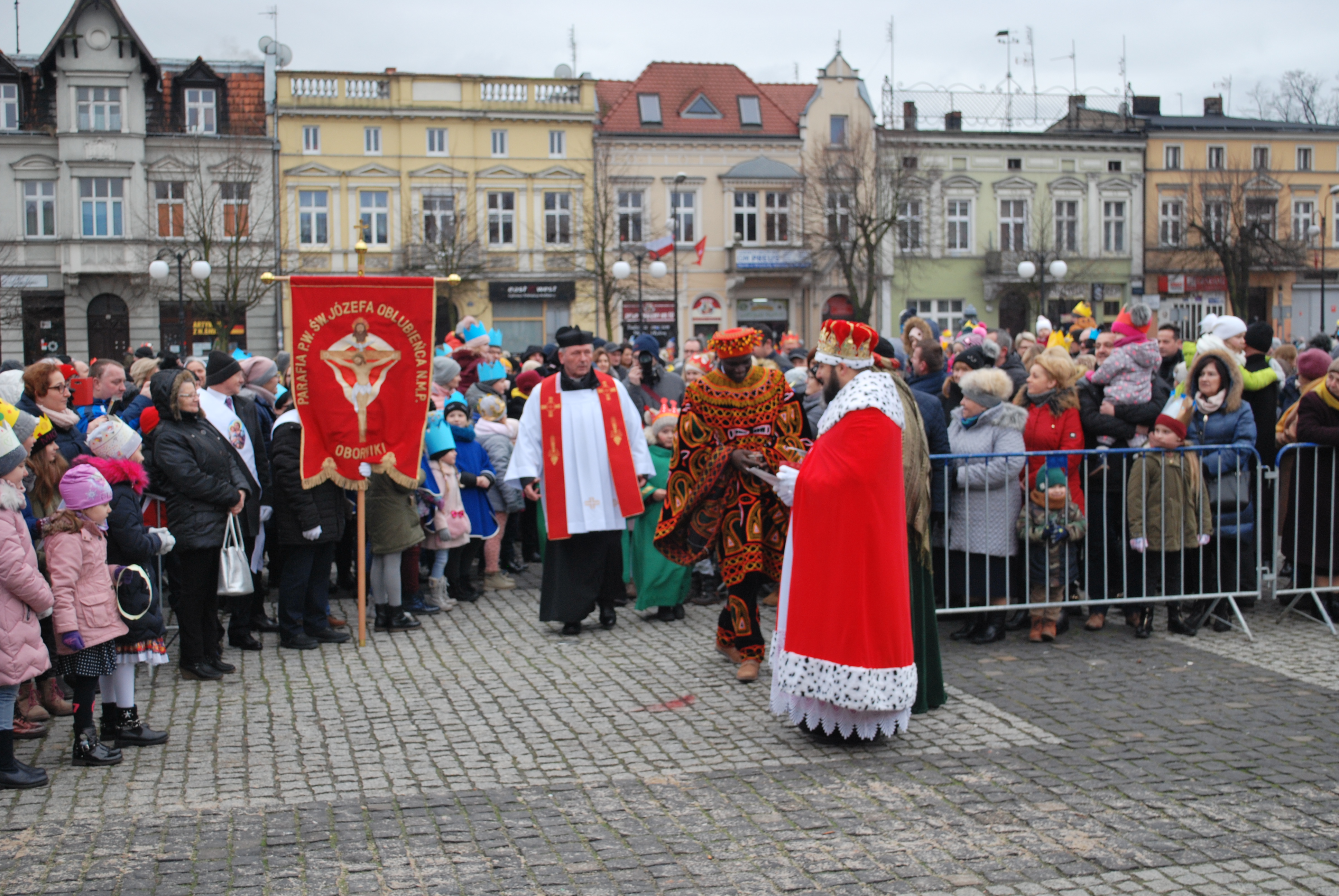 Korowód Trzech Króli przechodzi przez plac, otoczony budynkami i zgromadzoną publicznością.