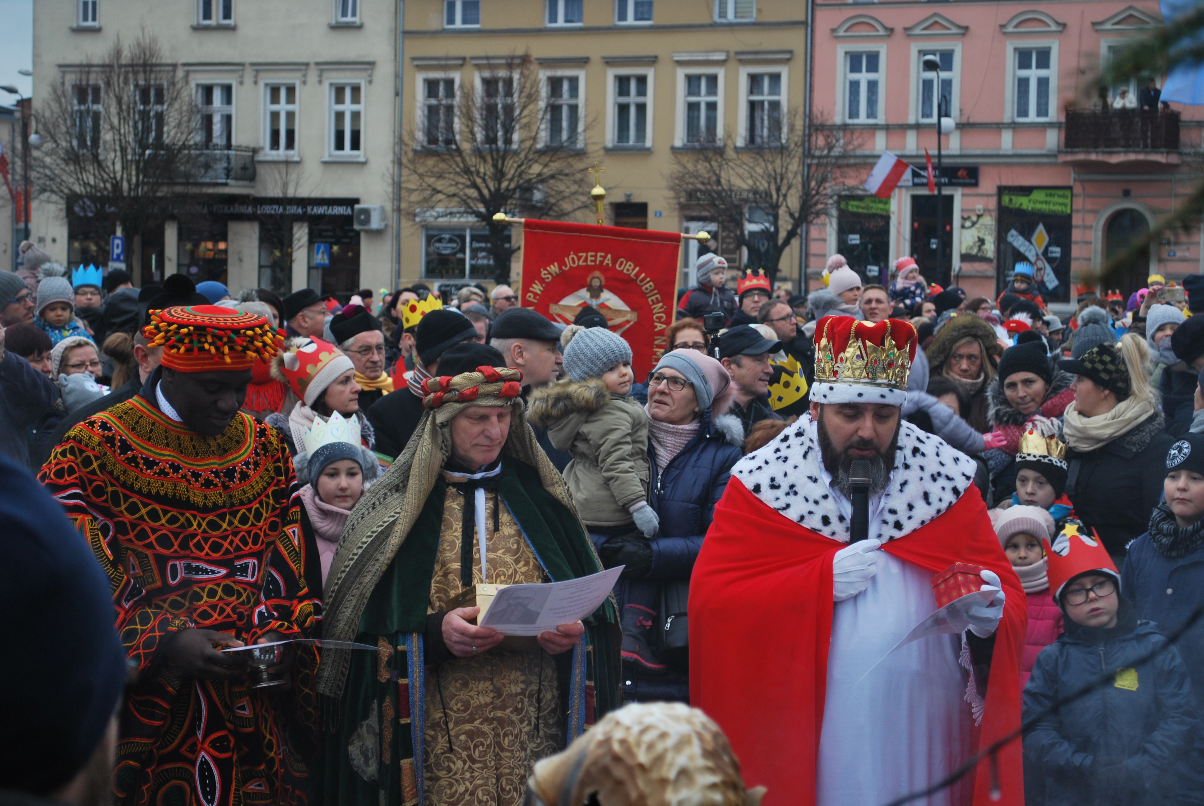 Korowód Trzech Króli w tłumie, uczestnicy w barwnych strojach i koronach, tło z budynkami.