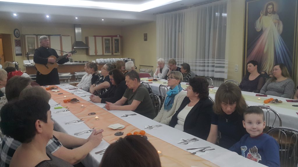 Spotkanie wigilijne pracowników: ksiądz gra na gitarze, uczestnicy siedzą przy nakrytych stołach